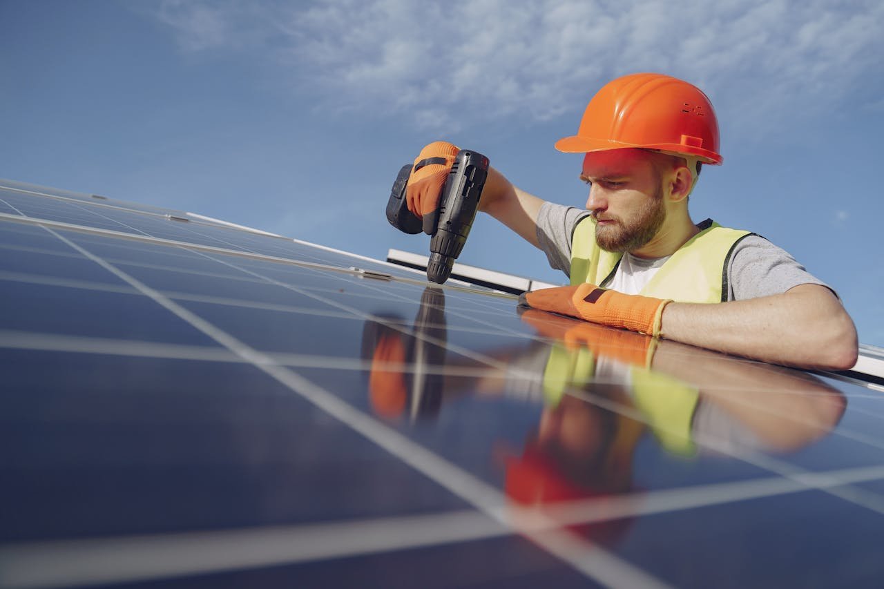 Low angle of serious bearded male electrician in hardhat and protective gloves installing solar photovoltaic panel system using drill